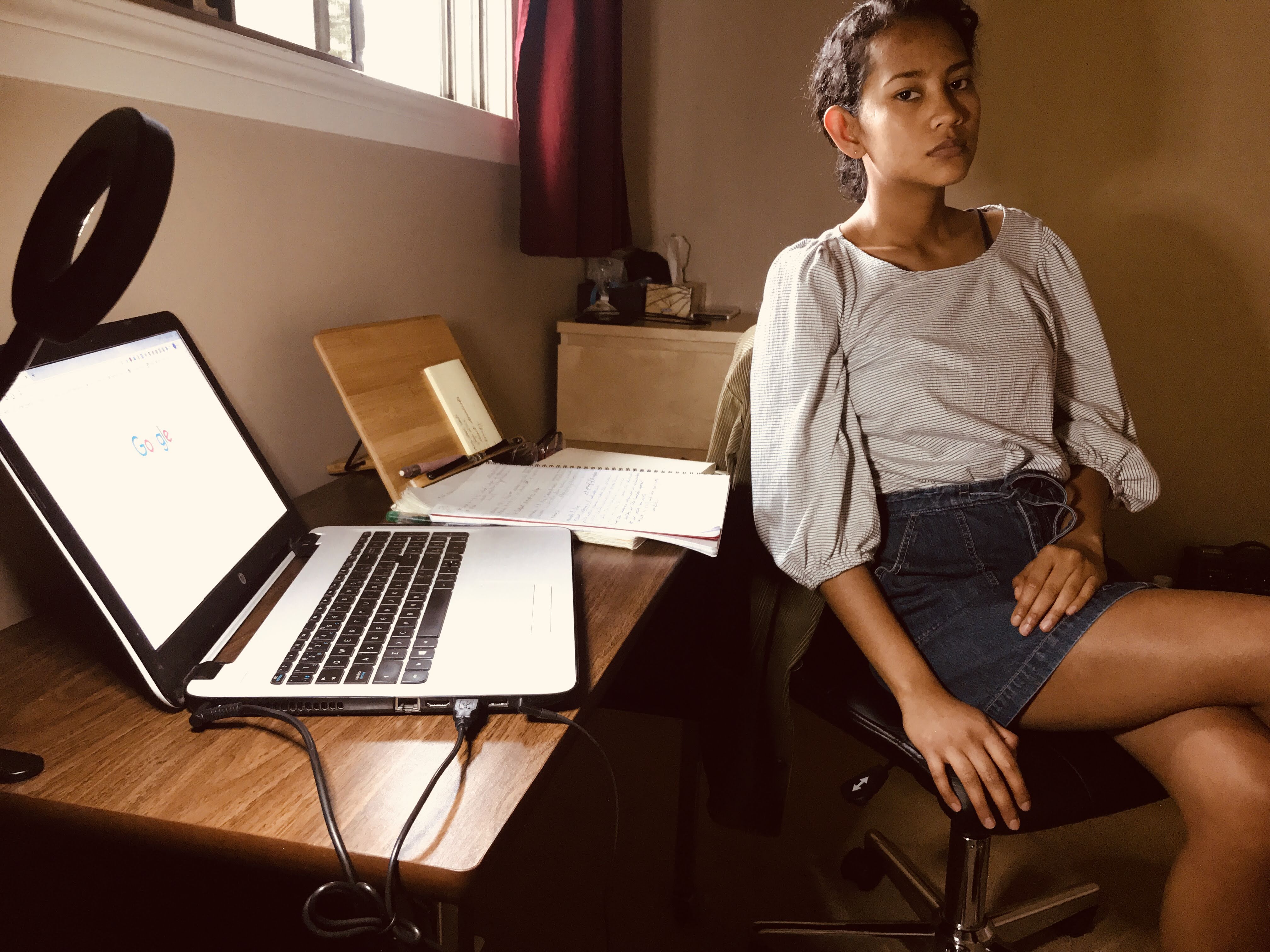 A photograph of a somber young woman in her room. She is at her desk and facing the camera. Her open laptop displays the Google homepage.