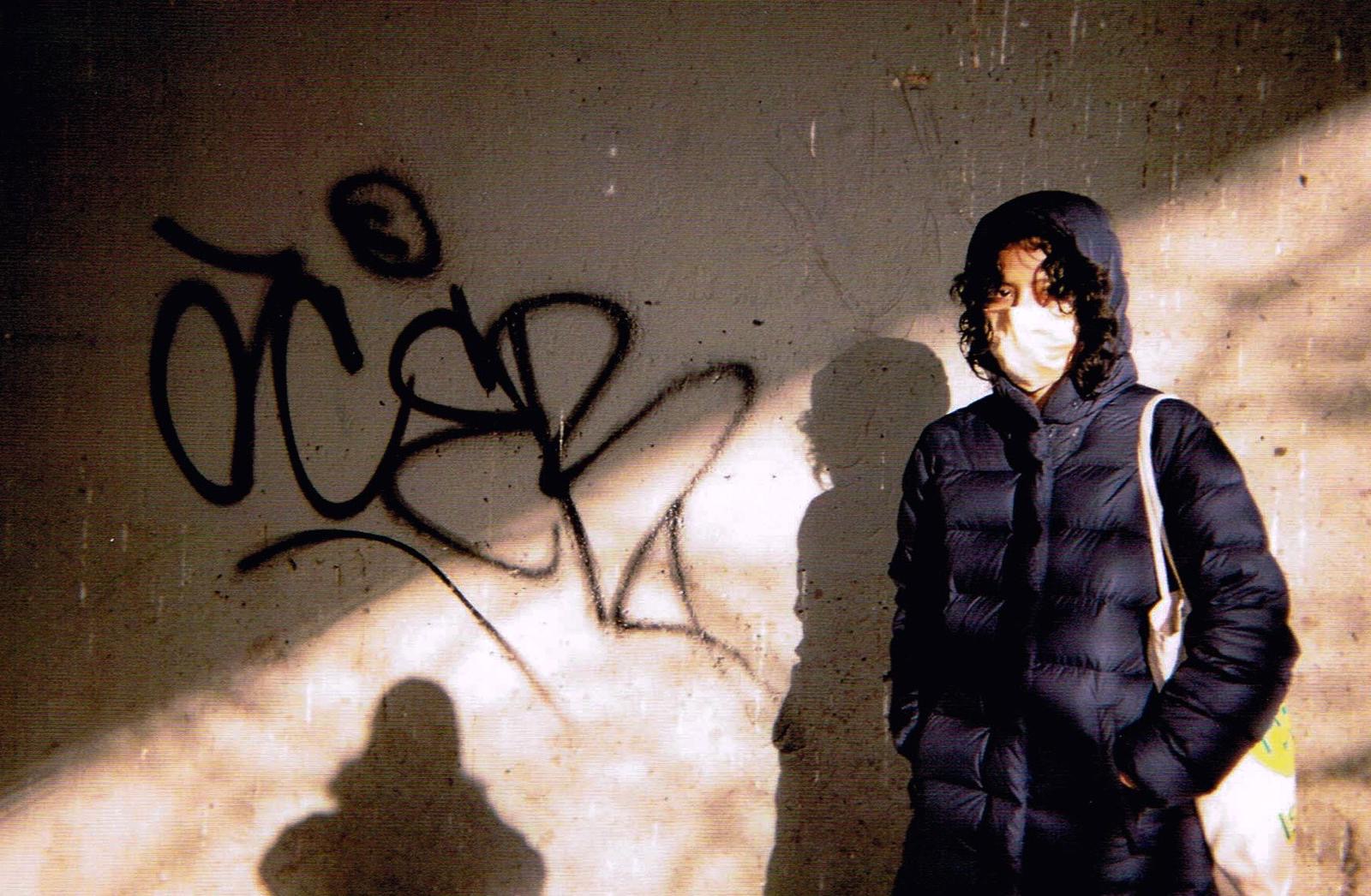 A photograph of a masked person in front of a graffitied cement wall. The photographer's shadow appears to the person's left.